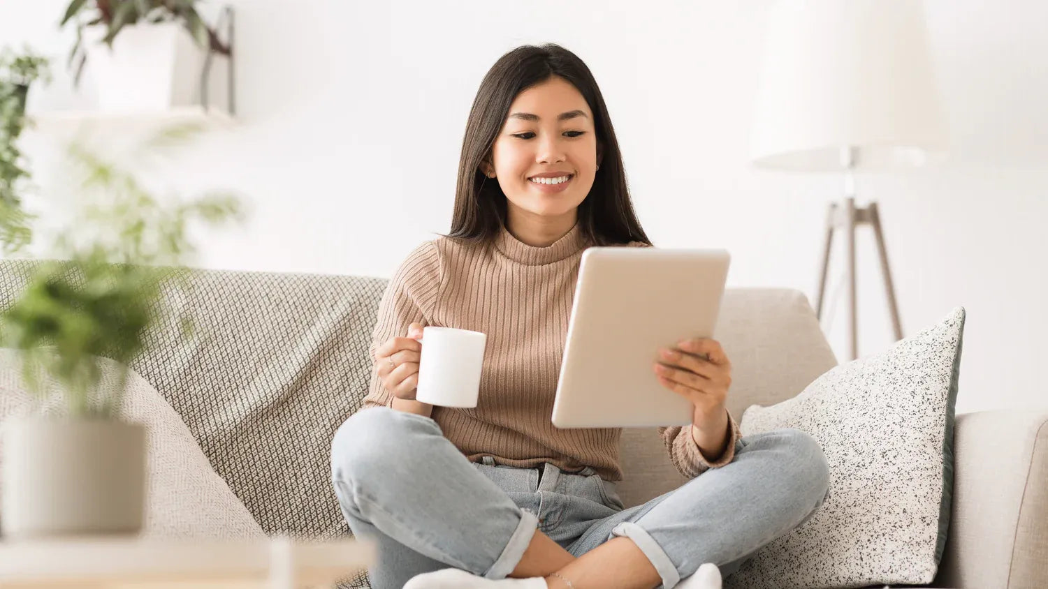 Eine junge Frau sitzt lächelnd mit einer Tasse Tee auf einem Sofa und liest in einem Tablet. Helle Lifestyle-Aufnahme als Banner für die Wissens-Kollektion mit Online-Kursen und E-Books.