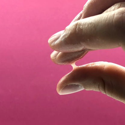 Imagen para determinar la fertilidad: se comprueba la capacidad de formar hilos del moco cervical entre el pulgar y el índice. El moco forma un hilo bien definido, lo que indica la fase de mayor fertilidad del ciclo menstrual. Fondo de color rosa.